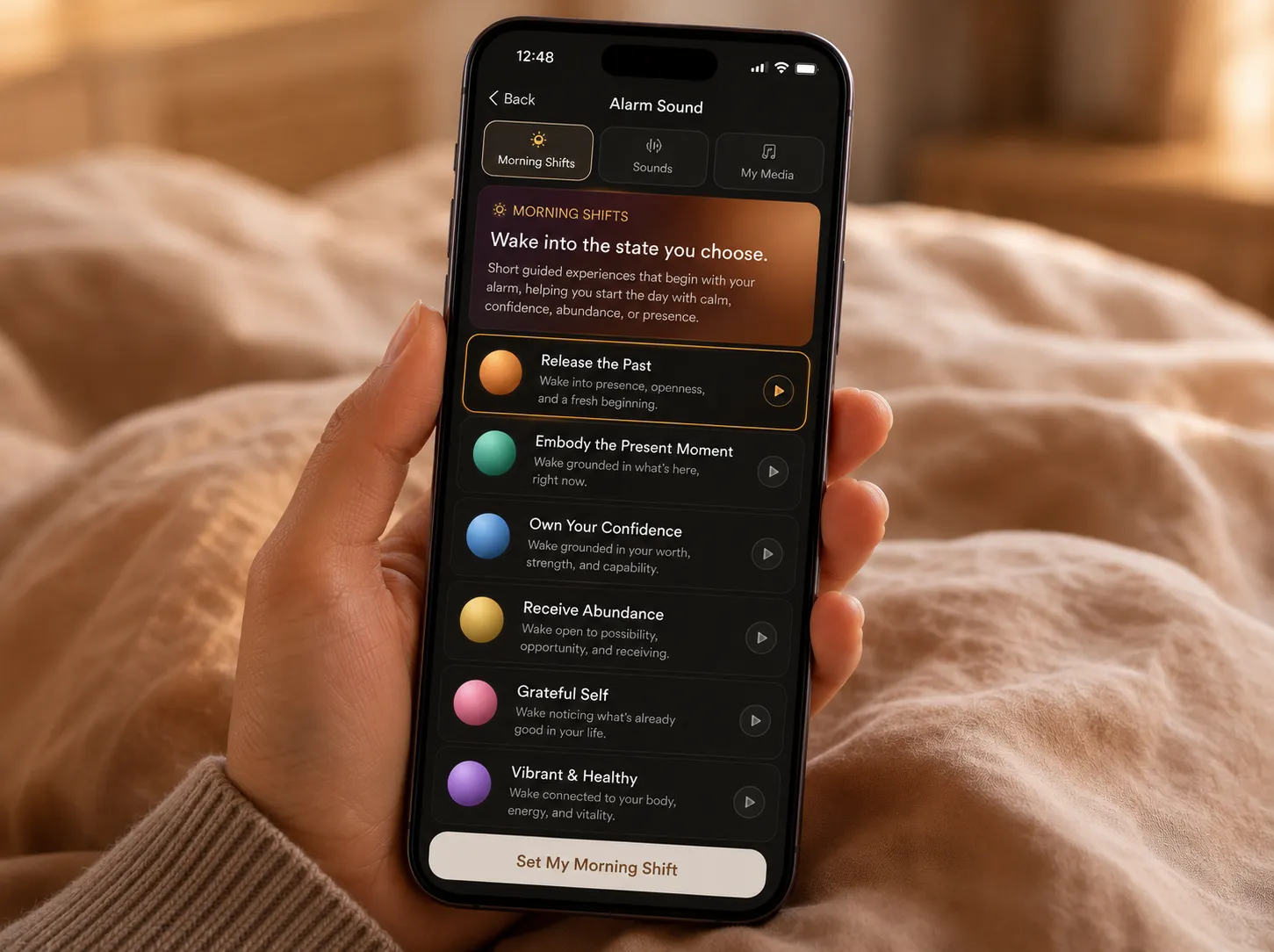 The Phoenix Rise Morning Shift picker held in one hand in bed, listing Release the Past, Embody the Present Moment, Own Your Confidence, Receive Abundance, Grateful Self and Vibrant & Healthy.