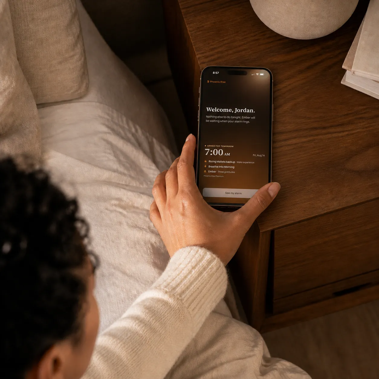 A woman reaching from bed toward a phone displaying her active Phoenix Rise morning alarm.