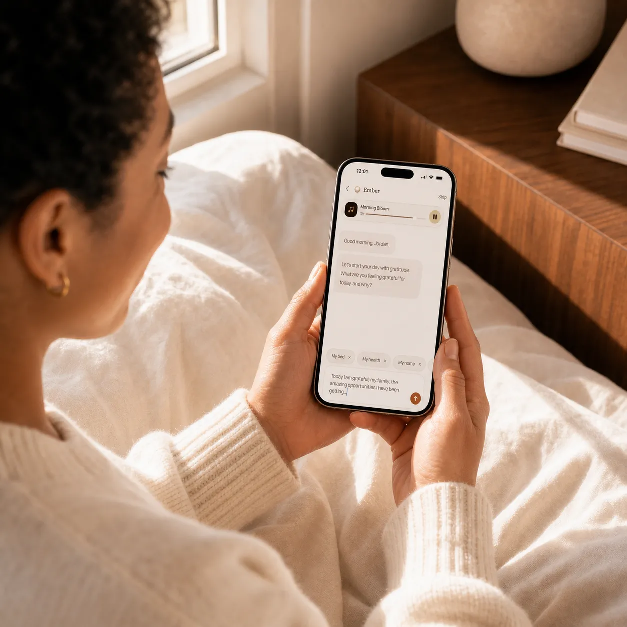 A woman sitting up in bed in warm morning light, responding to Ember’s guided gratitude prompt on her phone.